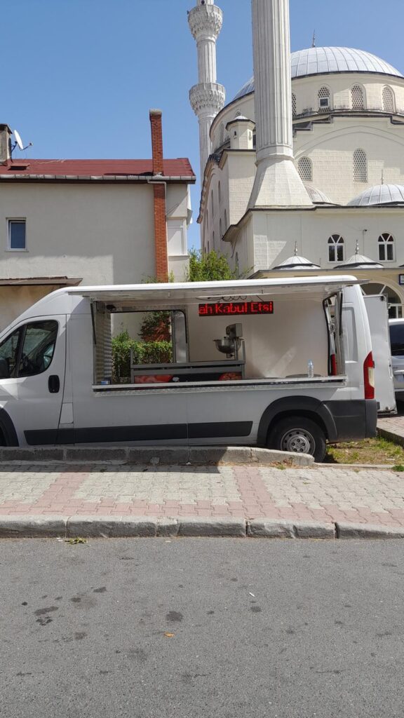Trafik içinde hayır lokması satılan mobil stand, cami ve eski binalar arka planda.